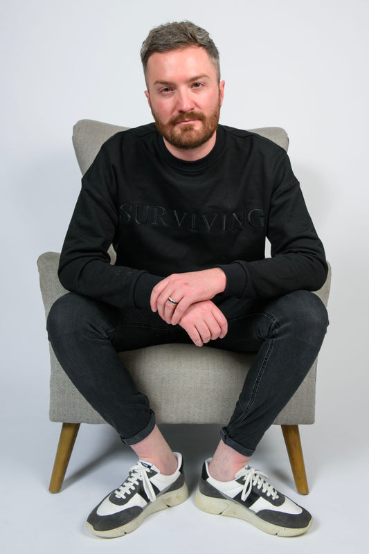 A man wearing the black Surviving mental health oversized unisex jumper sitting on an armchair 