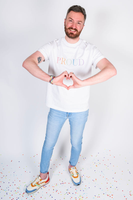 A beautiful white high quality oversized t-shirt with a pastel rainbow letters spelling out 'PROUD.' and the photo is showing the male making a heart shape with his hands