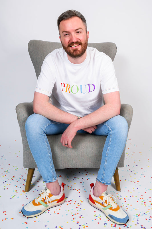A beautiful white high quality oversized t-shirt with a Bright rainbow letters spelling out 'PROUD.' and the photo is showing the male siting on an armchair wearing the t-shirt