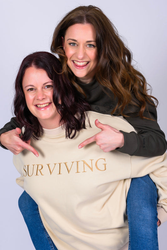 A lady wearing the cream Surviving mental health oversized unisex jumper giving a piggy back to her friend who is in the I am Enough jumper pointing at the Surviving writing