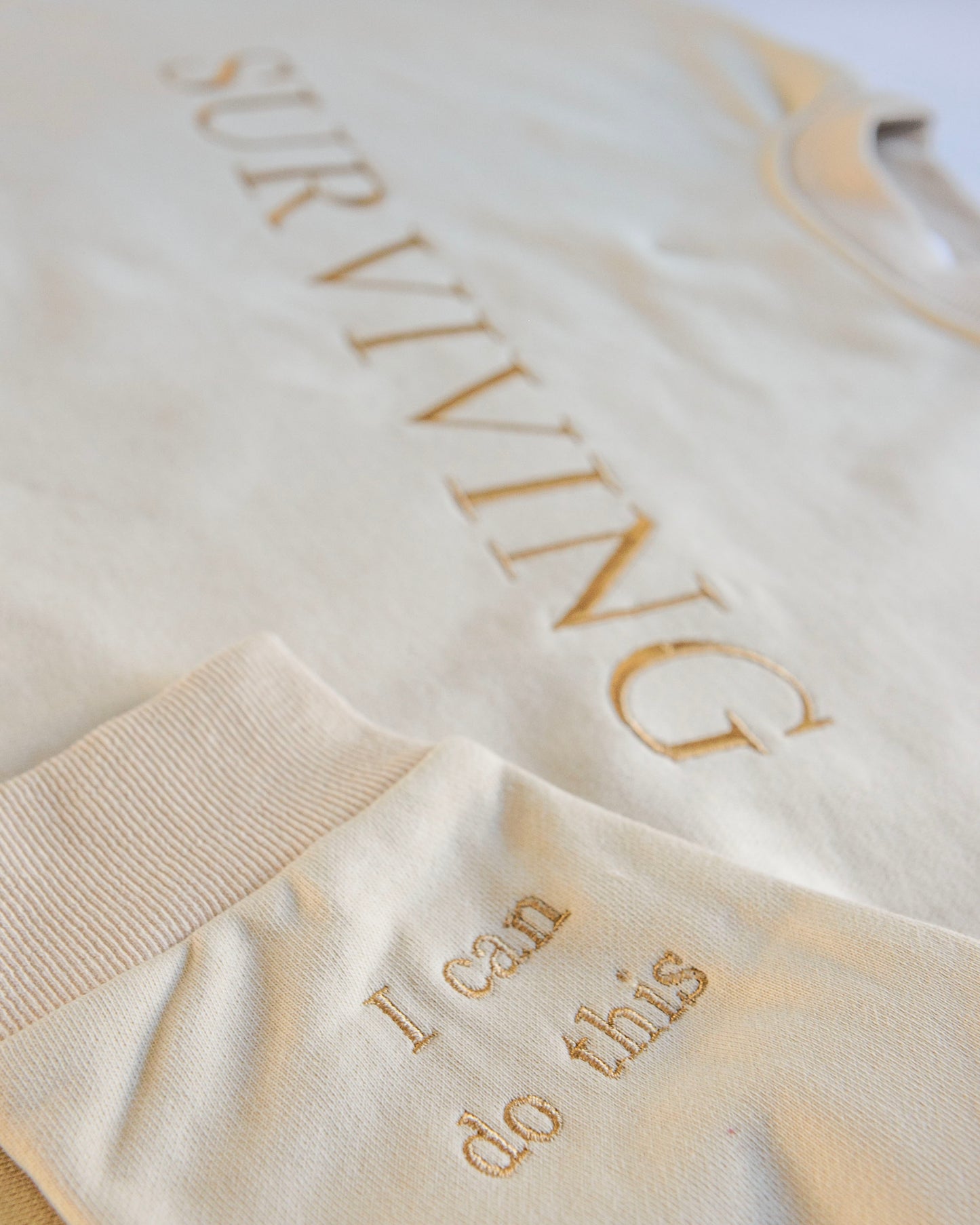 A beige, cotton oversized jumper with beautiful champaign embroidery it reads 'Surviving' in large capital letters across the chest and has reassuring messages on the cuffs that read 'I can do this' and 'Tomorrow's a new day '