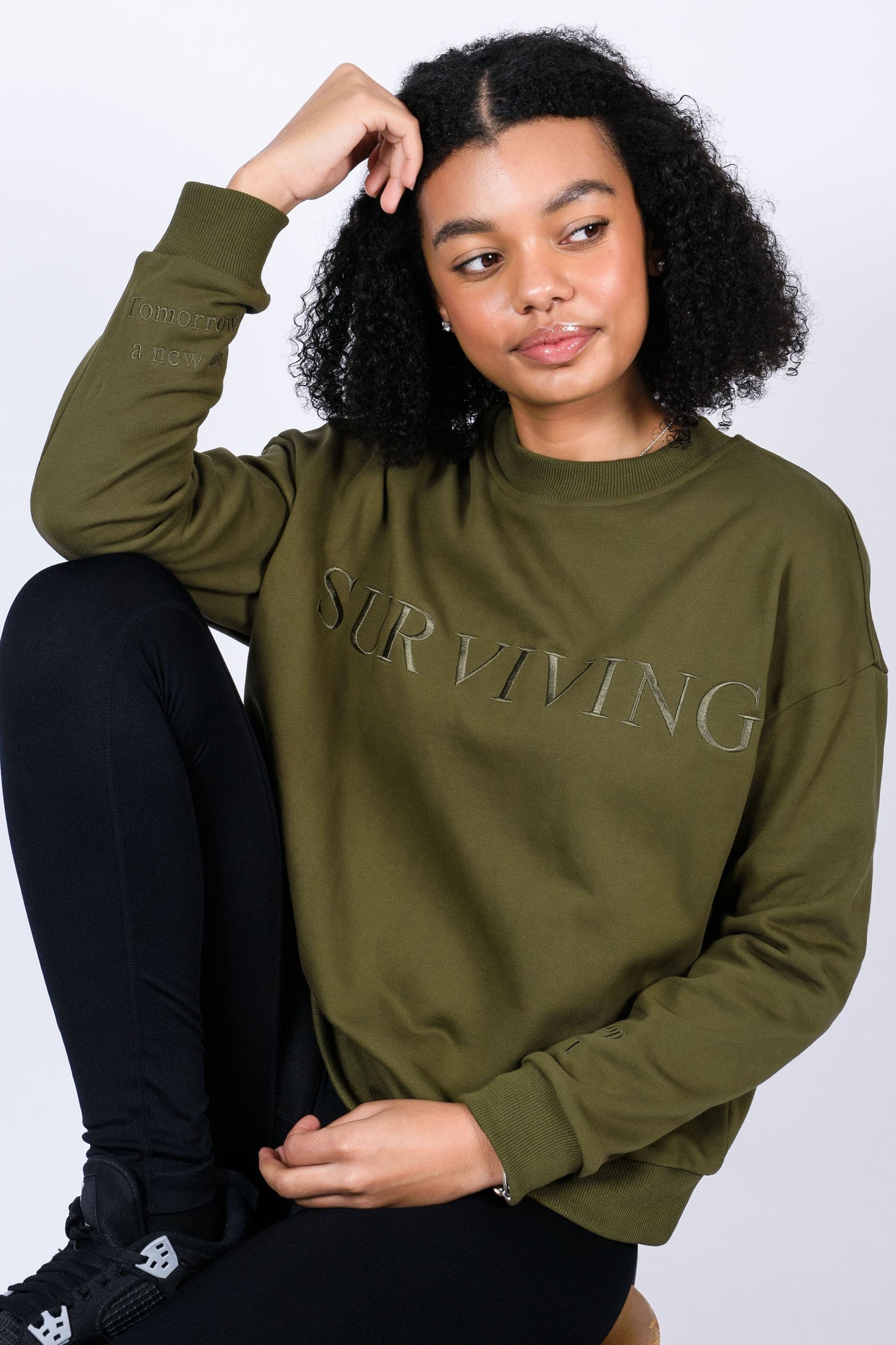 Our Surviving khaki green embroidered jumper from our mental health clothing collection, featuring the uplifting message "Surviving" across the chest. Additional encouraging phrases include "Tomorrow's a new day" and "I can do this," elegantly stitched to inspire resilience.