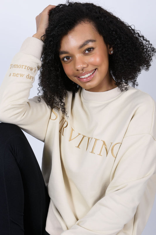 Our Surviving beige embroidered jumper from our mental health clothing collection, featuring the uplifting message "Surviving" across the chest and phrases include "Tomorrow's a new day" elegantly stitched to inspire resilience.