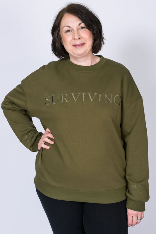 Our Surviving khaki green embroidered jumper from our mental health clothing collection, featuring the uplifting message "Surviving" across the chest. Additional encouraging phrases include "Tomorrow's a new day" and "I can do this," elegantly stitched to inspire resilience.